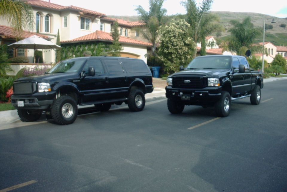 Two lifted black Ford trucks, an SUV and a crew-cab pickup, parked on a street