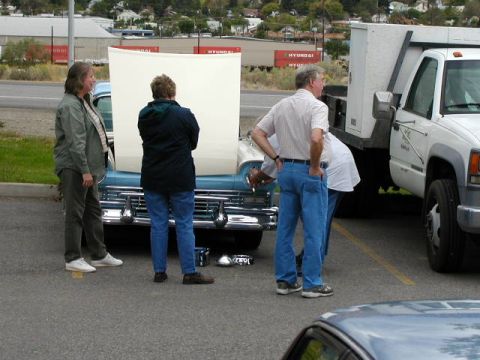 1967 Ford Ranchero - People inspect a vintage blue car with its hood open, parked beside a white utility truck.