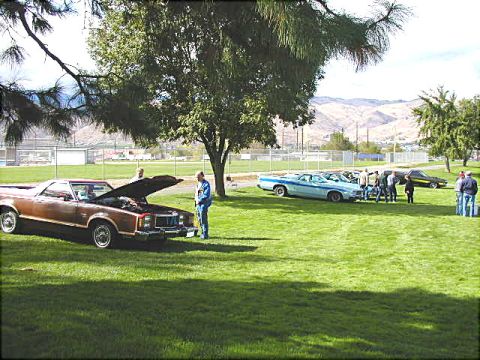 1967 Ford Ranchero - Several vehicles are parked on a grassy field under trees, with mountains in the background.