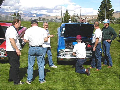 1967 Ford Ranchero - Several men examine the engine of a blue car, with another vehicle nearby on a grassy field under a cloudy sky.