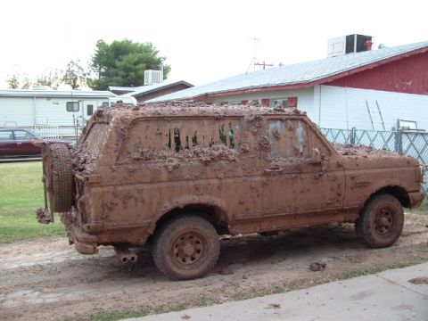 1990 Ford Bronco - This heavily mud-caked SUV, likely a 1987 Ford Bronco, is parked on a dirt surface in front of a house.