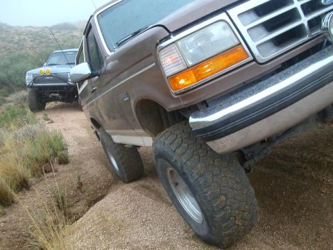 1993 Ford Bronco - Two Ford Broncos navigate a dusty trail in a hilly, overcast landscape.