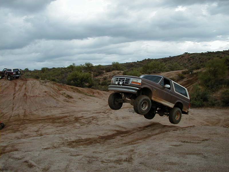 Brown Ford Bronco mid-air over a dirt mound, another SUV on a hill in background