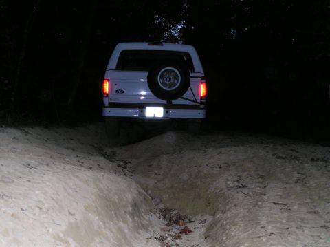 1996 Ford Bronco - A white 1996 Ford Bronco navigates a dirt trail in the dark, its taillights illuminated.