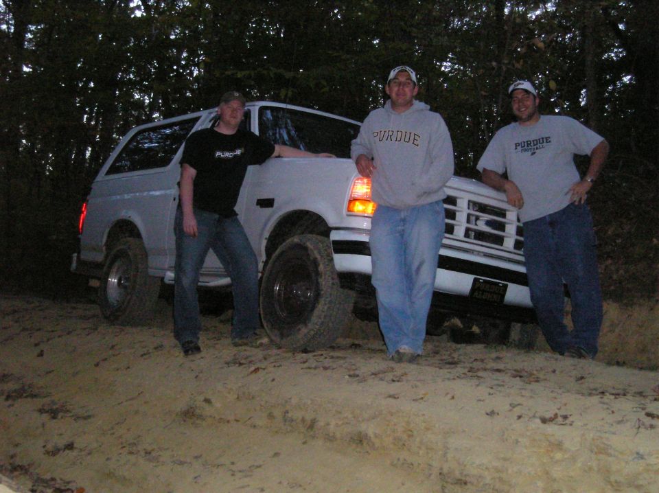 Three guys stand around a white Ford Bronco on a dirt path in the woods