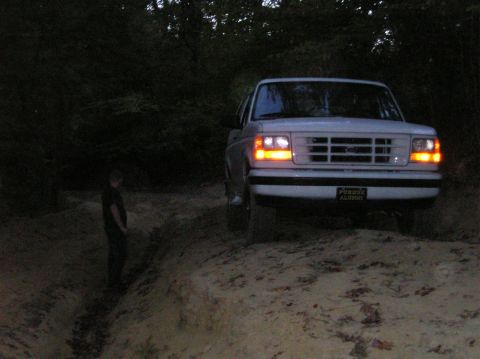 1996 Ford Bronco - A white Ford Bronco is seen from the front, its headlights illuminated, on a dirt path at dusk.