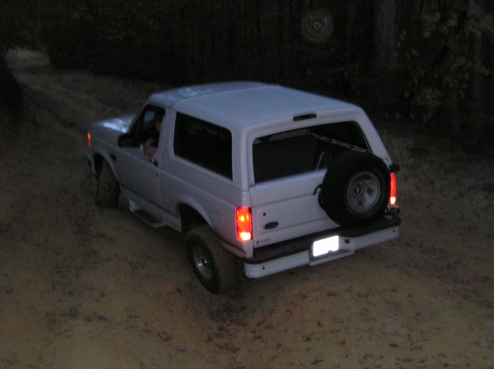 White Ford Bronco driving on a dirt road at dusk. Spare tire mounted on the back