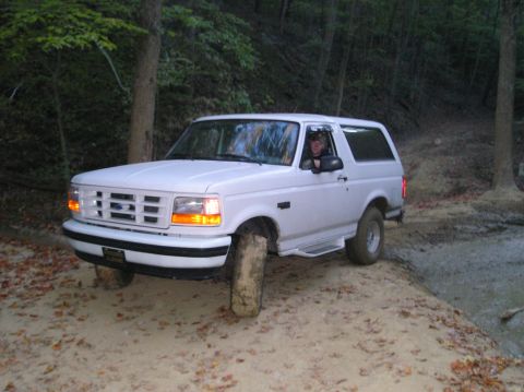 1996 Ford Bronco - A white Ford Bronco navigates a muddy off-road trail, with trees and foliage in the background.