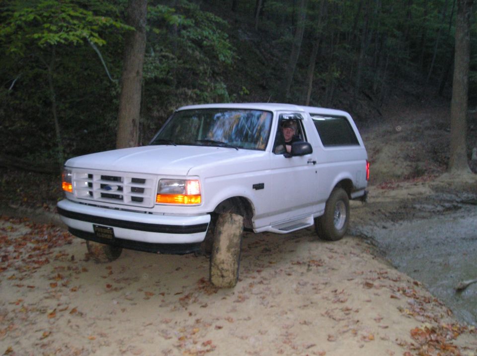 White Ford Bronco driving on a dirt trail with trees around. Driver is visible