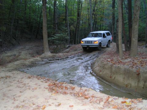 1996 Ford Bronco - A white 1996 Ford Bronco navigates a challenging muddy path in a wooded area.