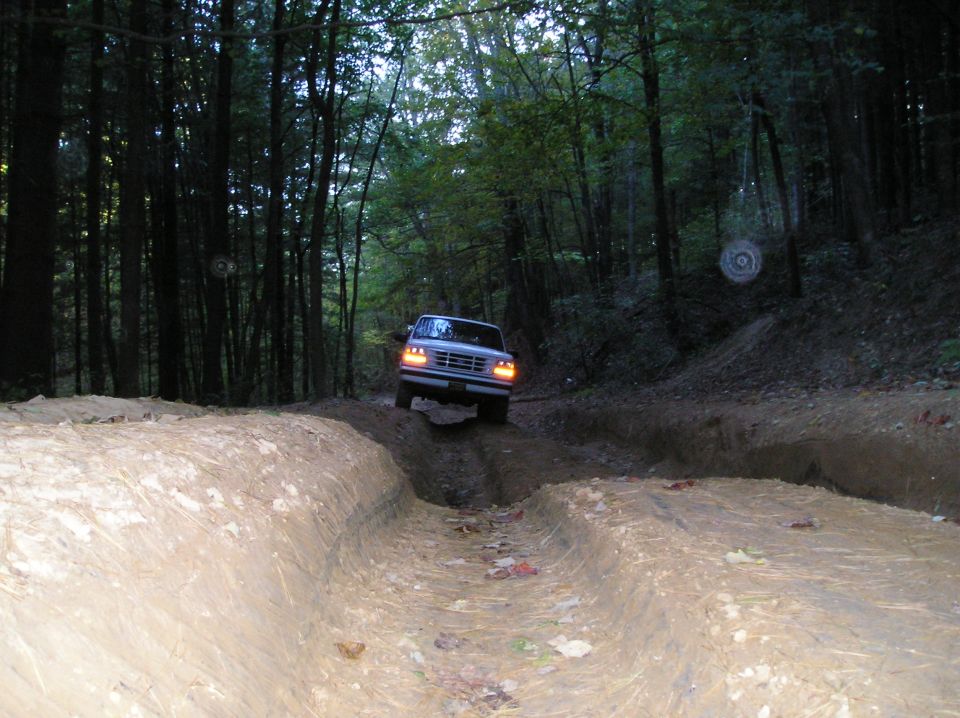 Ford Bronco driving through a muddy rutted trail in a wooded area. Headlights are on