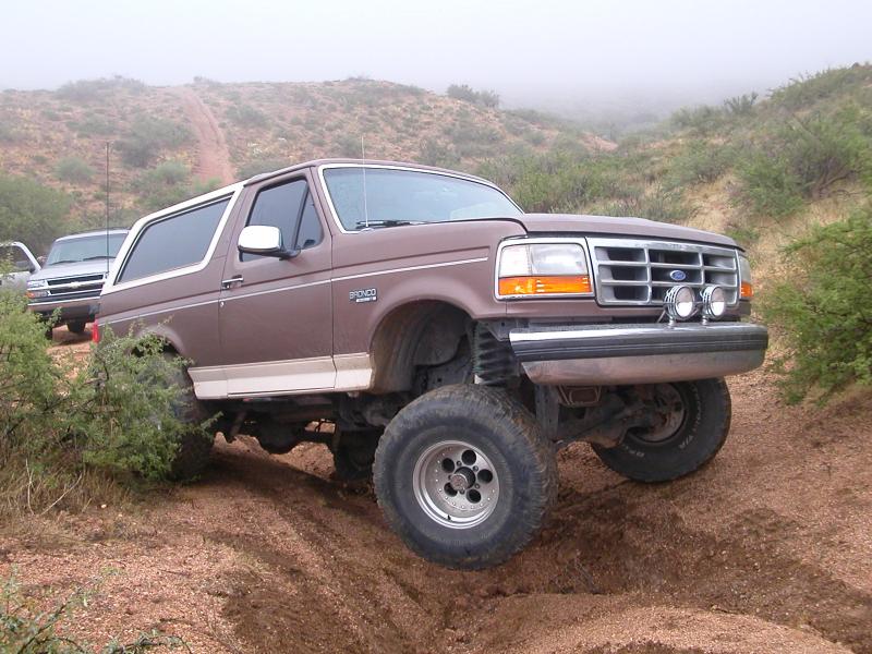 Brown Ford Bronco with lifted suspension driving on a dirt trail with another vehicle behind it