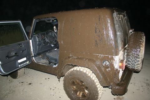 2003 Jeep TJ - A mud-caked 2003 Jeep TJ is viewed from the rear-right with its driver's door ajar.