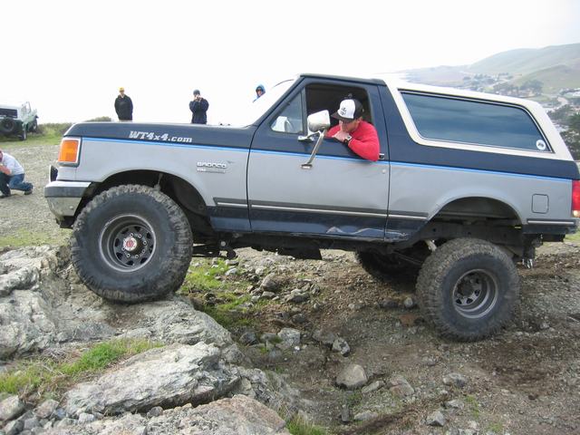 Lifted black-and-gray Ford Bronco flexing over rocks with driver leaning out window, side view