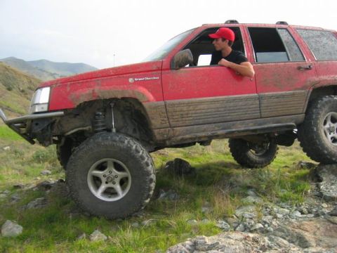 1989 Ford Bronco - A red Jeep Grand Cherokee navigates a rocky trail, its tires kicking up dirt.