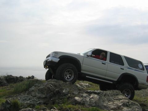 1989 Ford Bronco - A white SUV navigates a challenging rocky incline near the coast under an overcast sky.