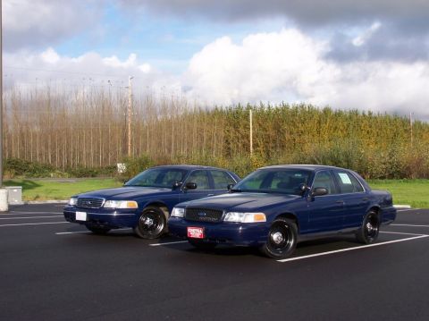1998 Ford Crown Victoria - Two dark blue sedans, likely police cruisers, are parked side-by-side in an outdoor lot under a cloudy sky.