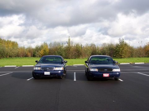 1998 Ford Crown Victoria - Two dark blue Ford Crown Victorias are parked facing forward in a parking lot under a cloudy sky.