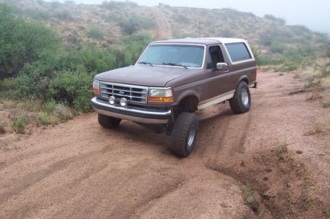 1993 Ford Bronco - A lifted brown Ford Bronco with a white top drives on a dusty, off-road trail.