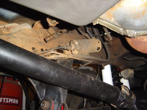 1986 Ford Bronco - Underbody shot looking up at a truck frame with a surface-rusted fuel tank and its mounting straps, hard fuel and brake lines, and a rusty…