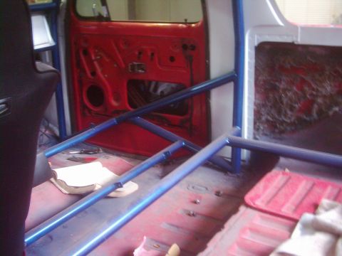 1978 Ford Bronco - The interior of a vehicle shows a blue roll cage, a red door, and a black racing seat, with a book on the floor.