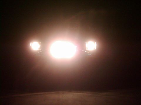 1978 Ford Bronco - The bright headlights of a vehicle illuminate the darkness, with a prominent central beam and two smaller side lights.