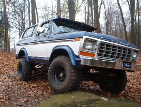 1978 Ford Bronco - A lifted 1978 Ford Bronco with a blue and white paint scheme sits on a rock in a wooded area.
