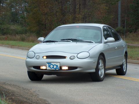 1998 Ford Taurus - Silver 1998 Ford Taurus driving on a paved road with trees in the background.