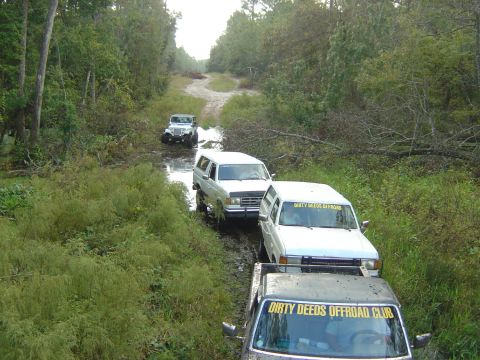 1993 Ford Bronco - A group of off-road vehicles navigate a wet and muddy trail surrounded by dense foliage.