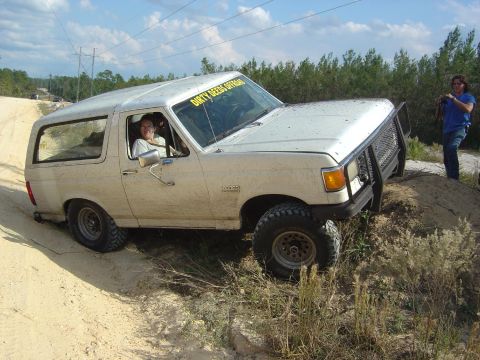 1993 Ford Bronco - A white Ford Bronco navigates an off-road trail, kicking up dust as it climbs a steep incline.