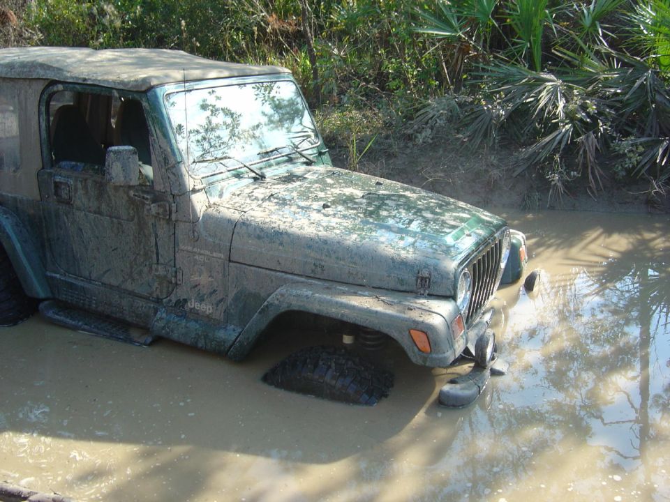 Mud-covered Jeep Wrangler stuck in a muddy ditch with vegetation in the background. Supermotors.net logo visible