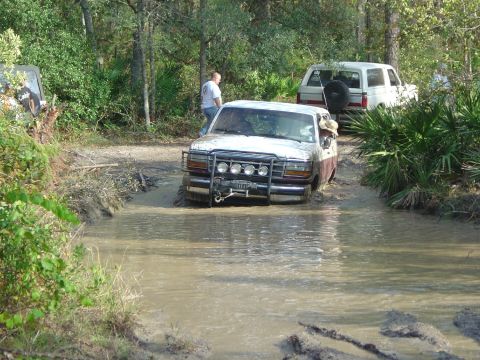 1993 Ford Bronco - A Ford F-Series truck navigates a muddy trail, with another SUV visible in the background.