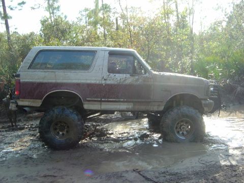 1993 Ford Bronco - This lifted Ford Bronco is navigating through a muddy off-road trail with trees in the background.