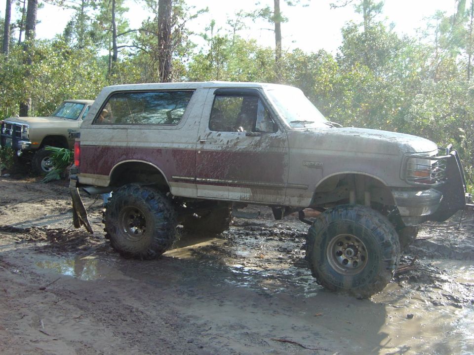 Mud-covered 1992 Ford Bronco with lifted suspension and off-road tires, parked in a muddy wooded area next to another truck