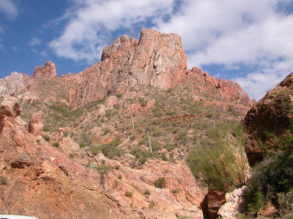 Rocky red mountain peak with scattered desert brush under a blue cloudy sky