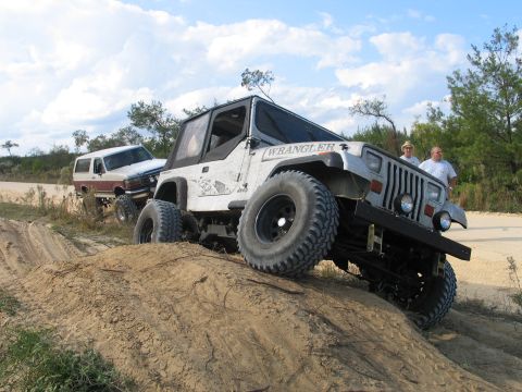 1992 Ford Bronco - A lifted white Jeep Wrangler YJ with a black soft top, black steel wheels and oversized mud-terrain tires flexes over a sand berm, one fron…