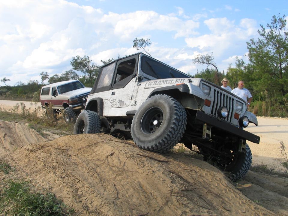 Lifted white Jeep Wrangler YJ flexing over a sand berm with a white and red Bronco behind