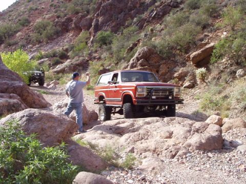 1993 Ford Bronco - A man directs a red Ford Bronco as it navigates a rough, rocky trail in a desert canyon.