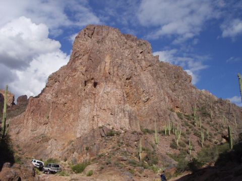 1993 Ford Bronco - Two SUVs are parked on a dirt path in a desert canyon, with a large rock formation and saguaro cacti under a blue sky.