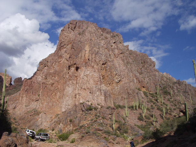 White SUV and silver truck parked below a huge saguaro-covered desert butte