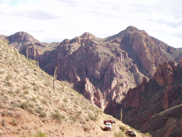 Two 4x4s on a narrow desert shelf road below rugged red rock ridges