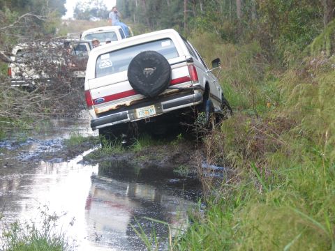 1992 Ford Bronco - Rear three-quarter view of a white and red two-tone Ford Bronco flexed at a steep angle up a muddy embankment beside a water-filled trail,…