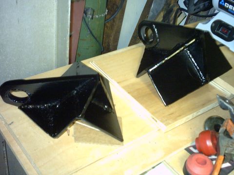 1995 Ford Bronco - A matched pair of freshly welded and gloss-black-painted fabricated suspension link brackets sitting on a plywood workbench, each with a gu…