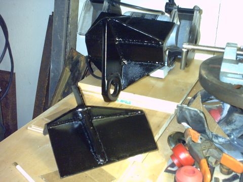 1995 Ford Bronco - Two shop-fabricated black-painted steel brackets with welded gussets sit on a plywood workbench, the upper one fitted with a large recovery…