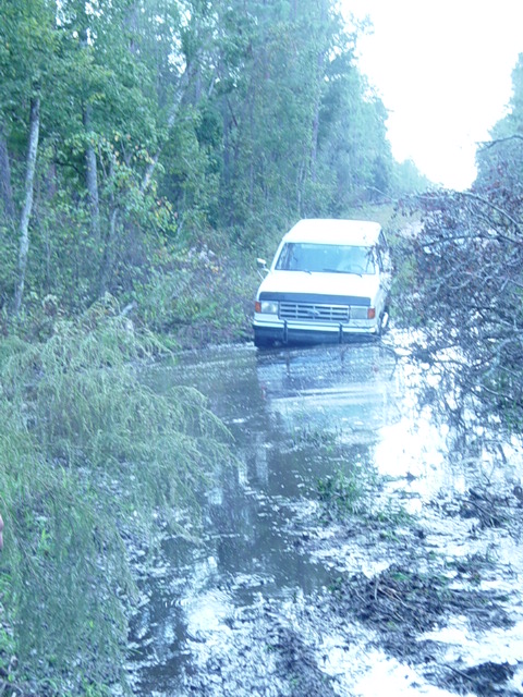 1989 Ford Bronco - A white van is stuck in a large puddle of muddy water on an unpaved road.