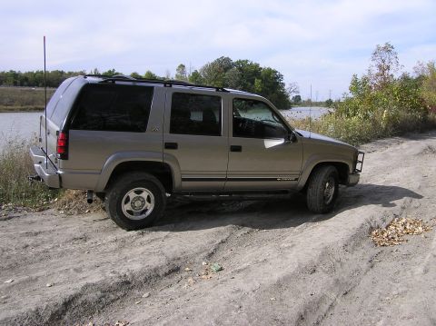 2000 Chevrolet Tahoe - A tan SUV is parked on a dirt road with a body of water and trees in the background.