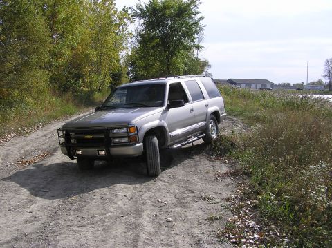 2000 Chevrolet Tahoe - A silver SUV with a brush guard is parked on a dirt road with trees and buildings in the background.