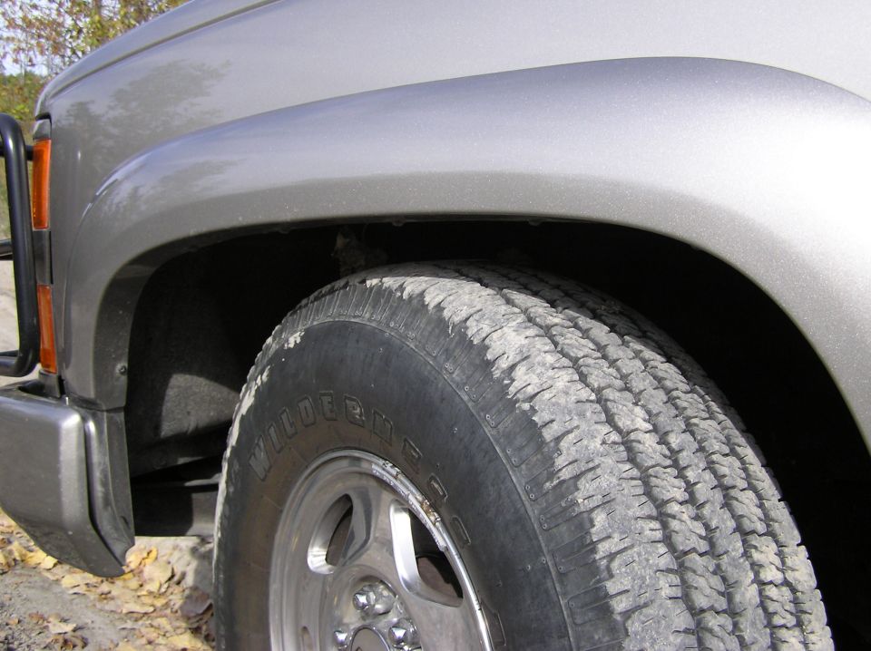 Close-up of a silver SUV's front fender and tire. The tire has 'WILDERME' visible