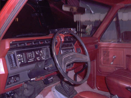 1986 Ford Bronco - The interior of a red truck shows the dashboard, steering wheel, and seats. The dashboard has square gauges and vents.