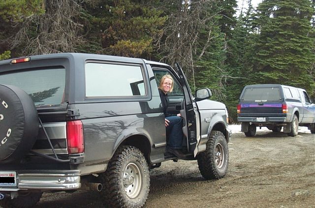 1995 Ford Bronco with woman in driver's seat, parked on dirt road with trees. Another truck is behind
